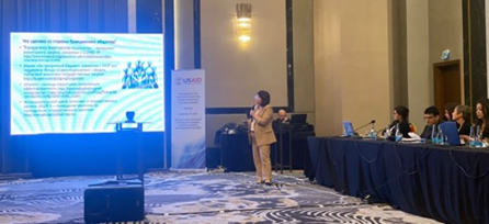 Woman with microphone in front of PowerPoint presentation and USAID banner